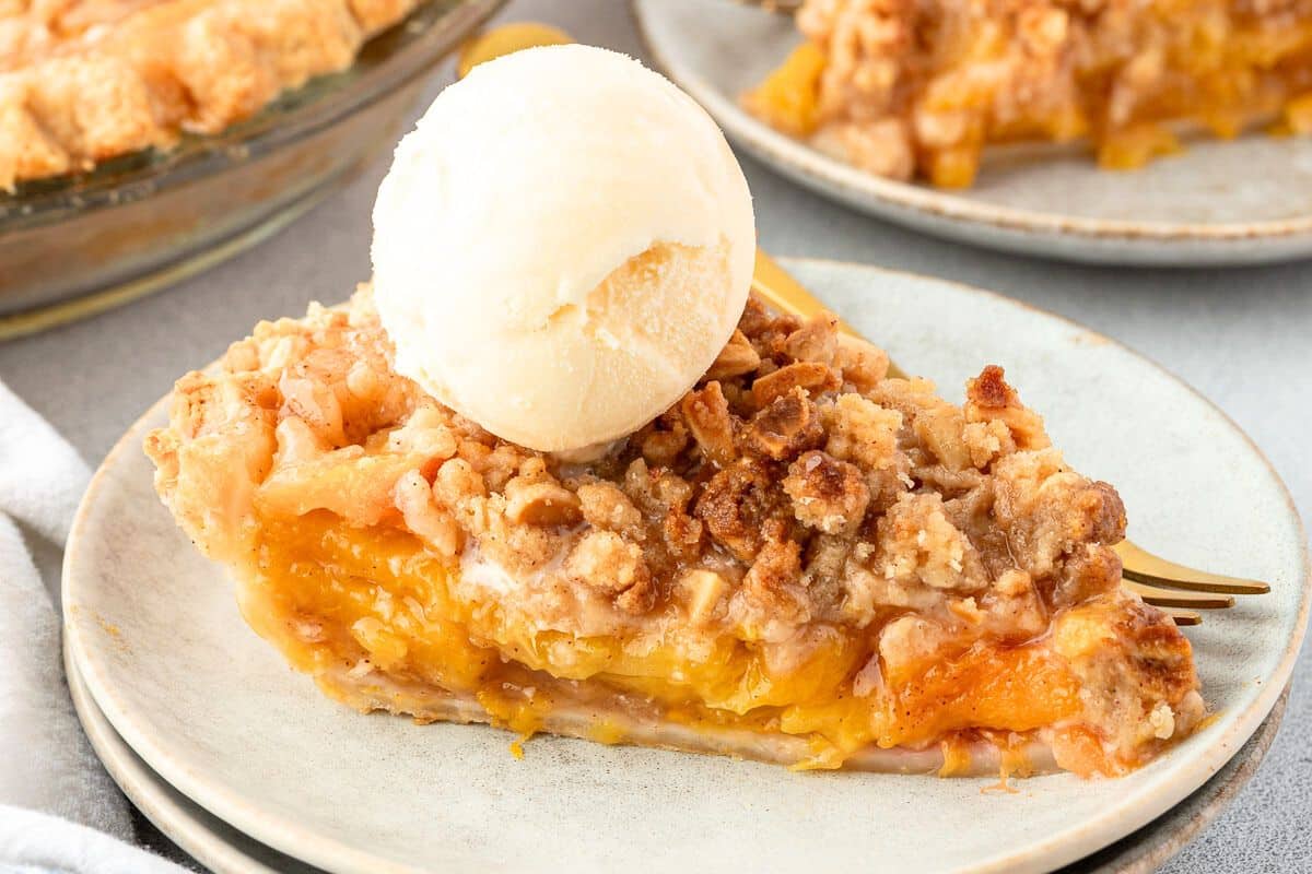 A slice of peach crumble pie topped with a scoop of vanilla ice cream on a plate with a fork.