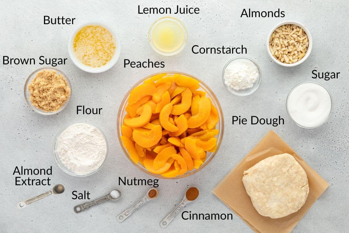 Ingredients for peach pie, including peaches, pie dough, and various labeled baking ingredients on a white surface.
