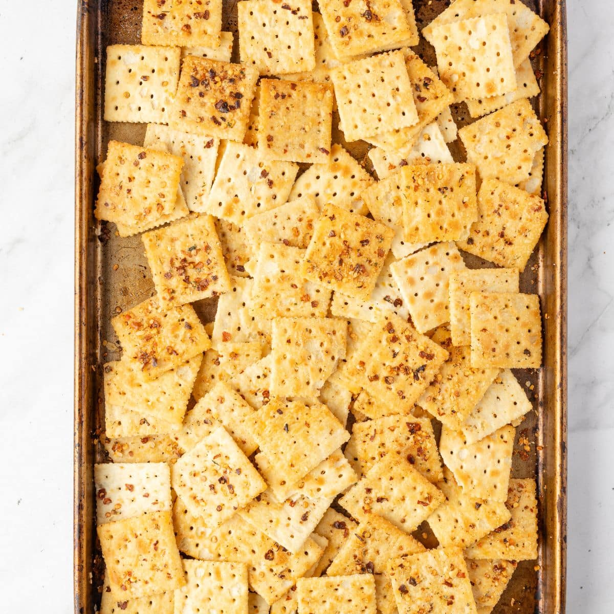 A baking sheet filled with seasoned square crackers, lightly dusted with spices.