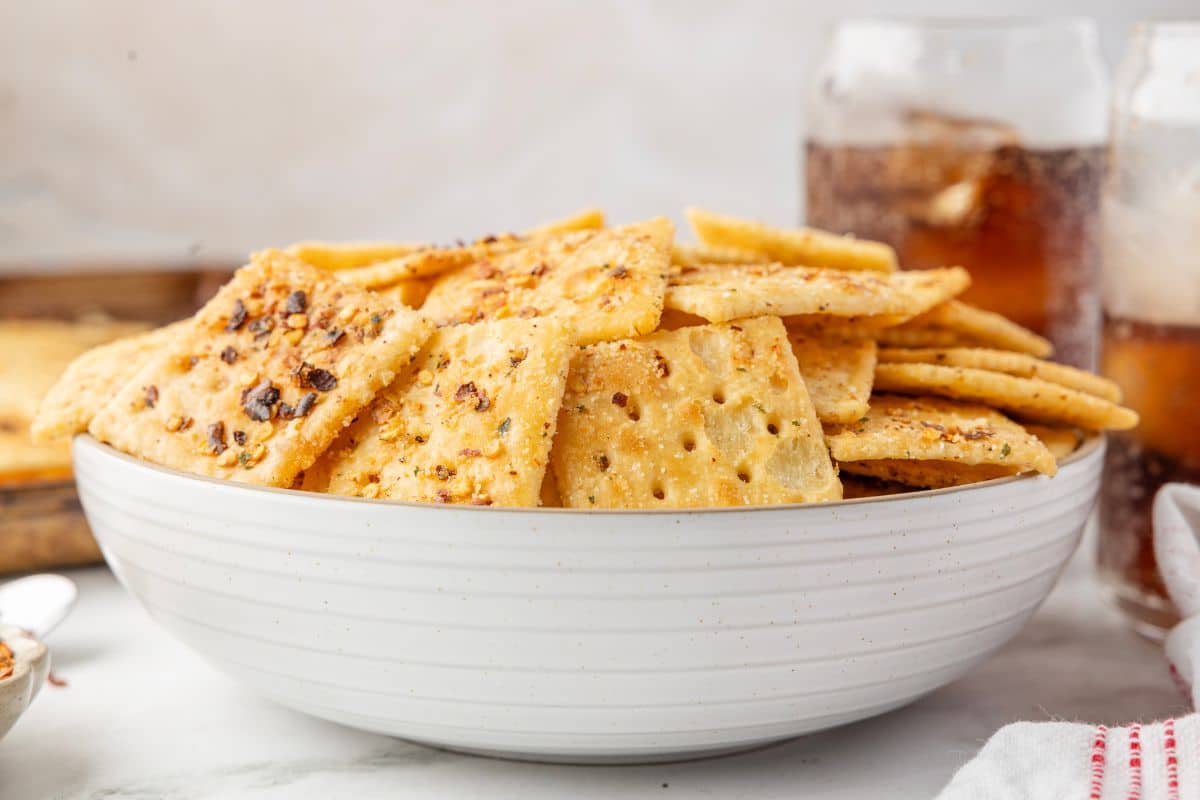 A white bowl filled with seasoned square crackers, with glasses of iced soda in the background.