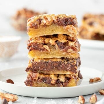 Three stacked dessert bars with chocolate chips and nuts on a white plate, with more bars in the background.
