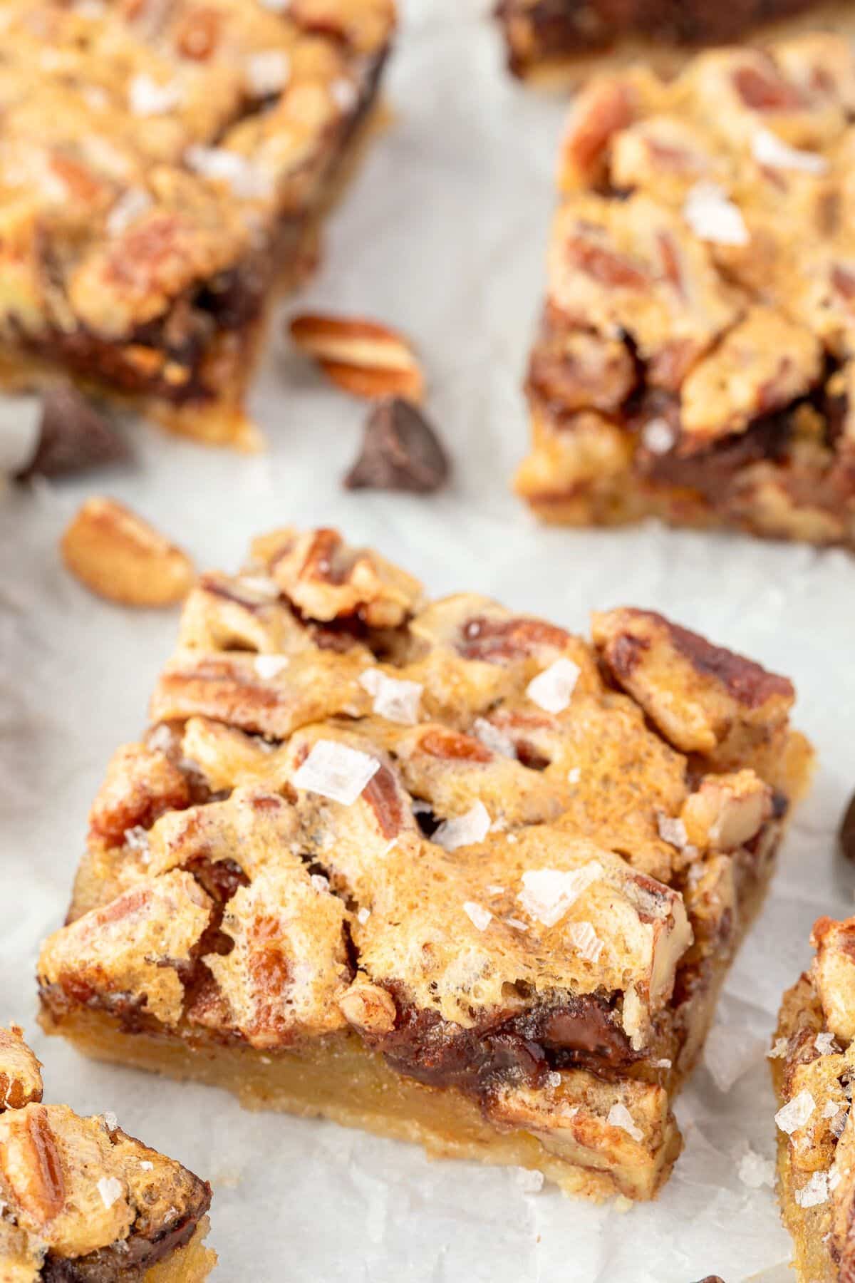 A close-up of a pecan chocolate chip dessert bar topped with flaky sea salt on parchment paper.