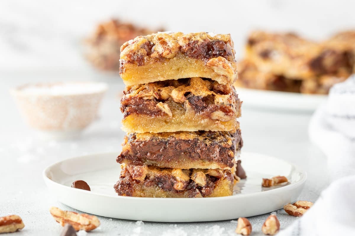 A stack of three pecan and chocolate dessert bars on a white plate, with nuts scattered around.