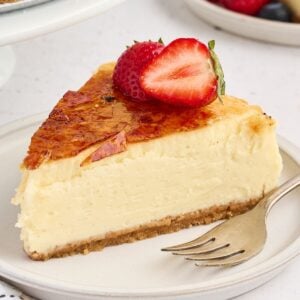 A slice of cheesecake with caramelized top, garnished with a halved strawberry, on a plate with a fork.