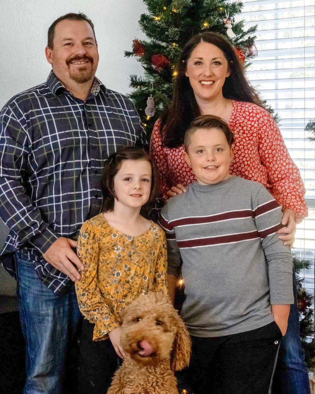 A smiling family with two kids and a dog posing in front of a decorated Christmas tree.