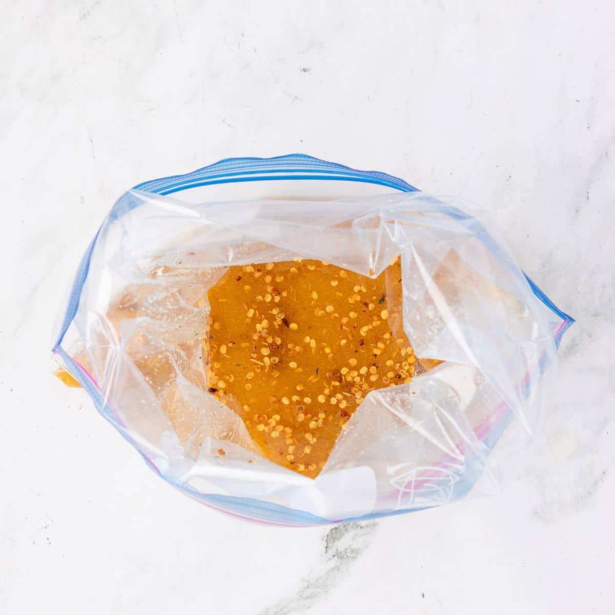 A plastic zip-top bag filled with orange marinade and red chili flakes on a white surface.