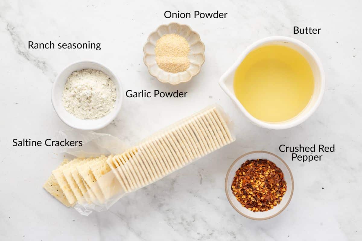 Saltine crackers, butter, ranch seasoning, garlic powder, onion powder, and crushed red pepper on a white surface.