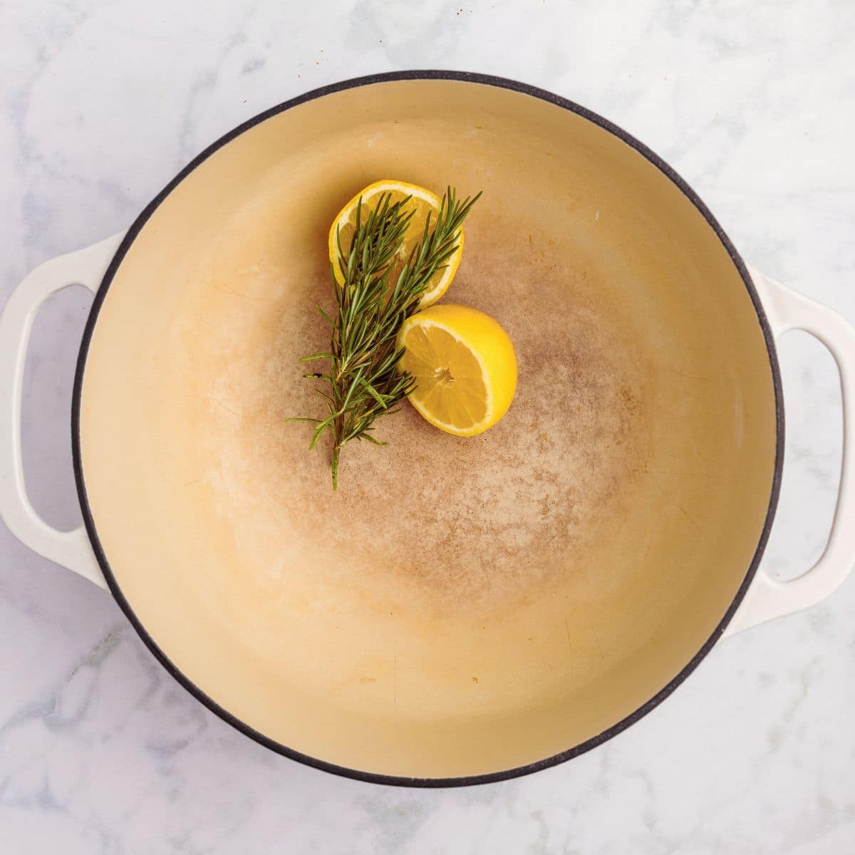 A white Dutch oven with two lemon halves and a sprig of rosemary inside on a marble surface.