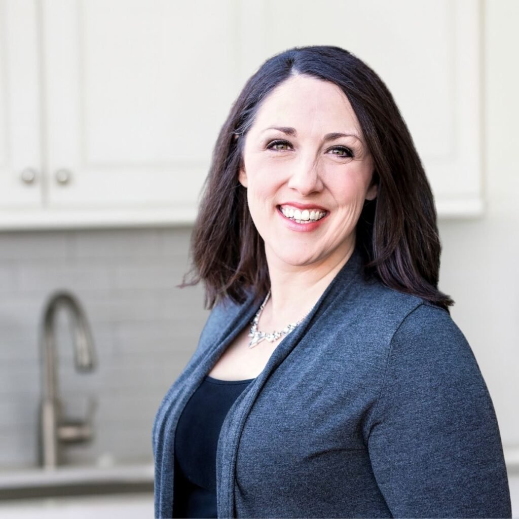 Woman with dark hair in a gray sweater smiling in a bright kitchen with white cabinets and a silver faucet.