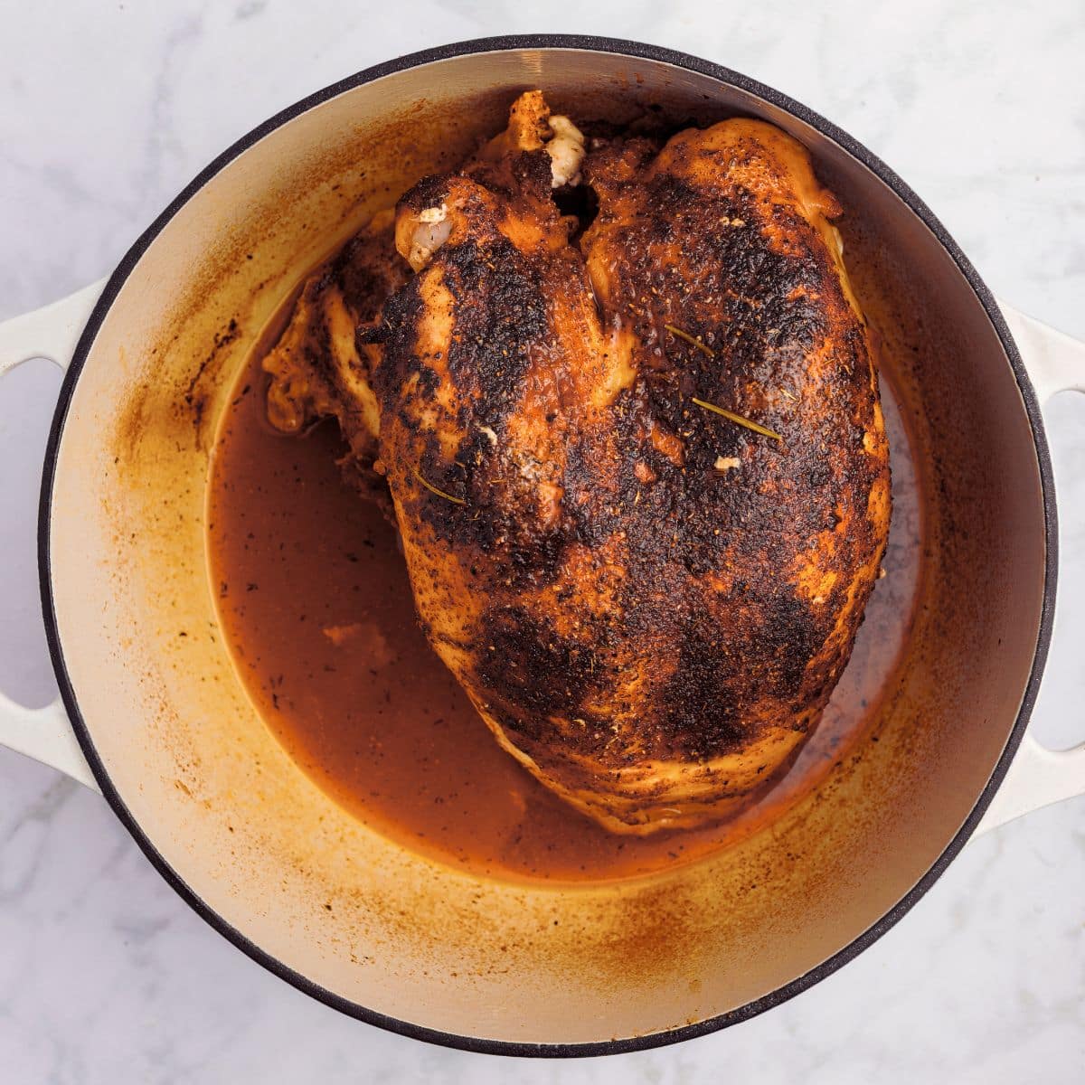 Roast chicken with crispy skin in a round pot with brown juices, viewed from above on a marble surface.