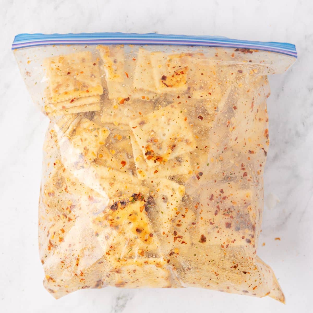 Plastic zipper bag filled with square crackers coated in red pepper flakes and seasoning, on a white surface.