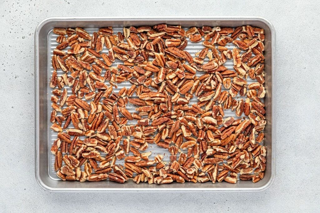 A baking tray with a single layer of chopped pecans spread out evenly on it.