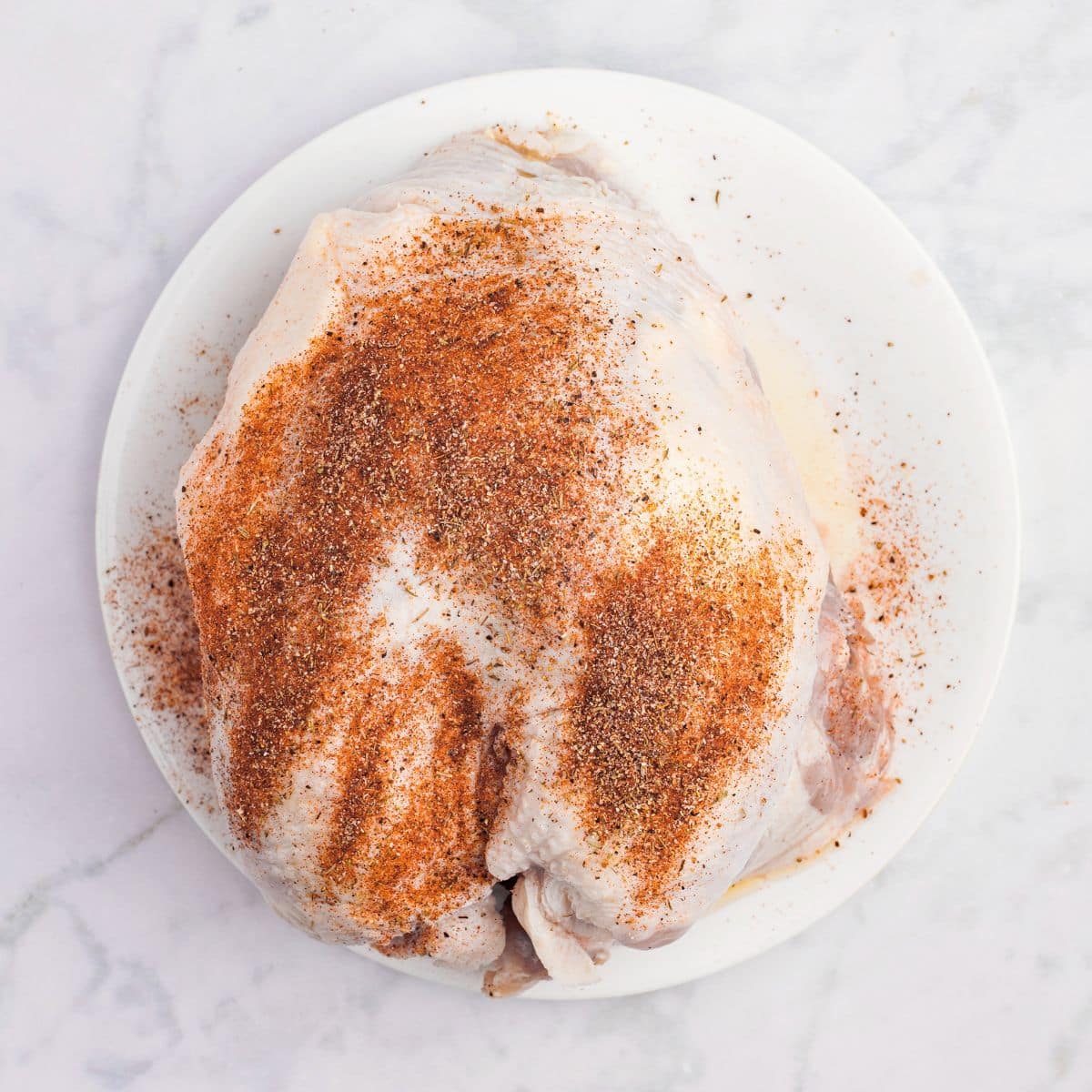 Raw turkey breast on a white plate, sprinkled with a reddish-brown spice rub, on a marble surface.