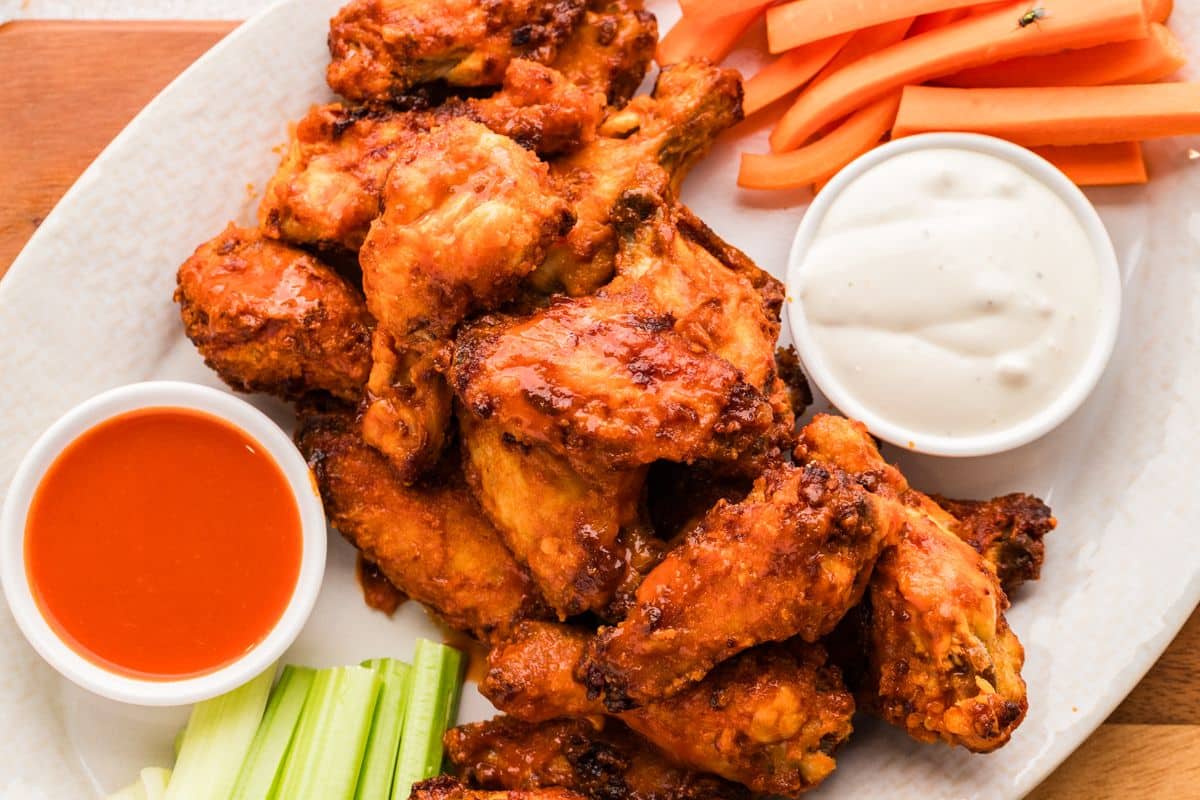 Plate of buffalo chicken wings with celery, carrot sticks, ranch dip, and hot sauce on the side.