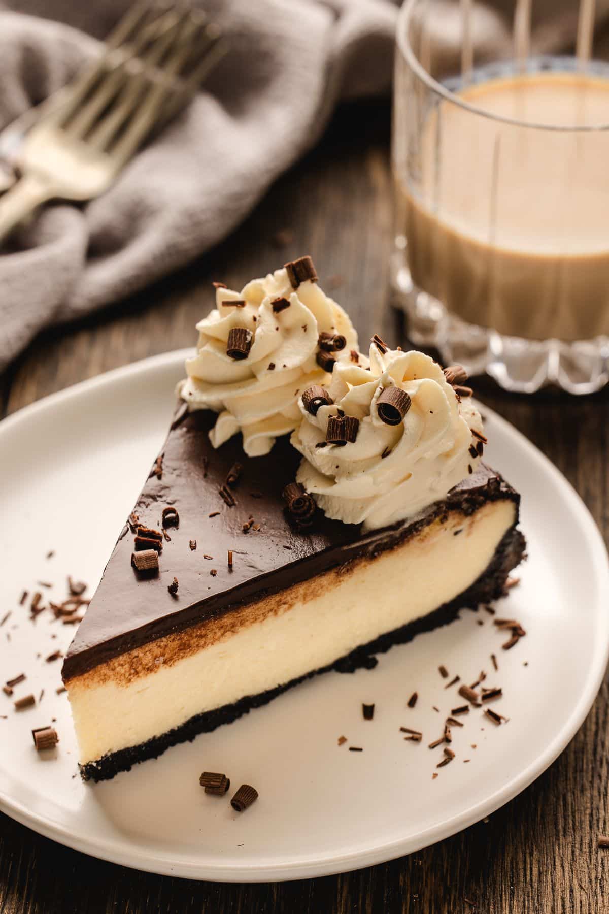 A slice of chocolate-topped cheesecake with whipped cream and chocolate shavings on a white plate.