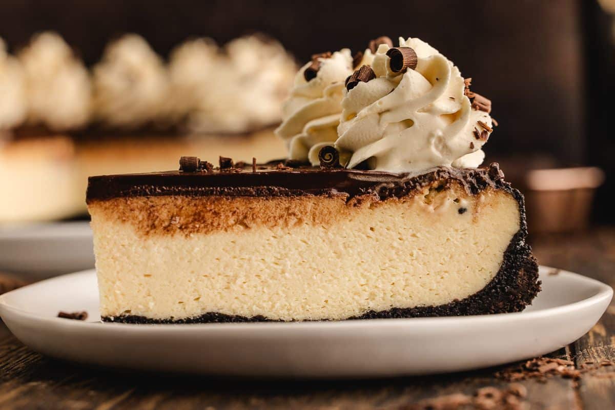 A slice of cheesecake with chocolate crust, chocolate topping, whipped cream, and chocolate curls on a plate.
