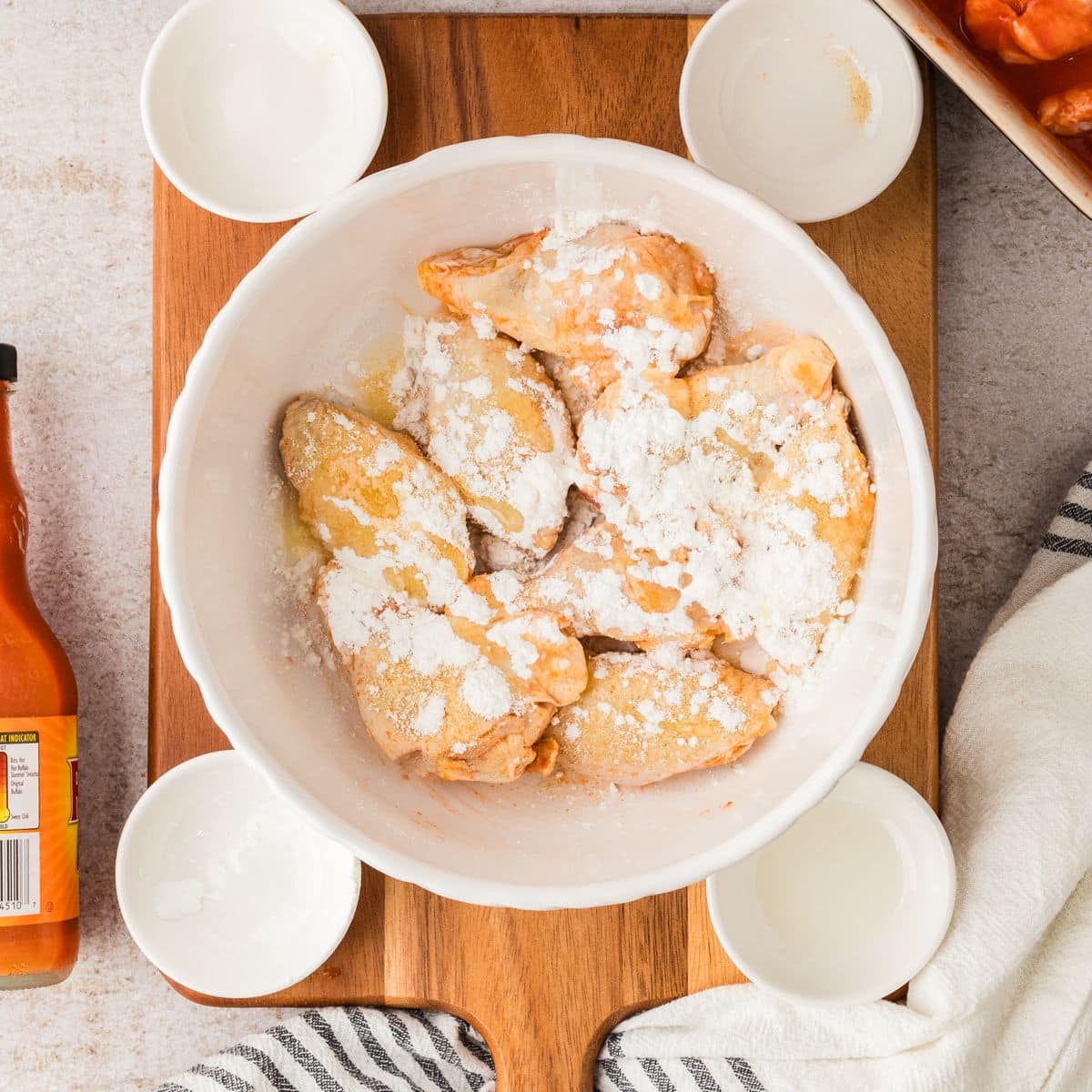 Raw chicken pieces coated with baking soda in a white bowl, surrounded by small bowls and a bottle of hot sauce.