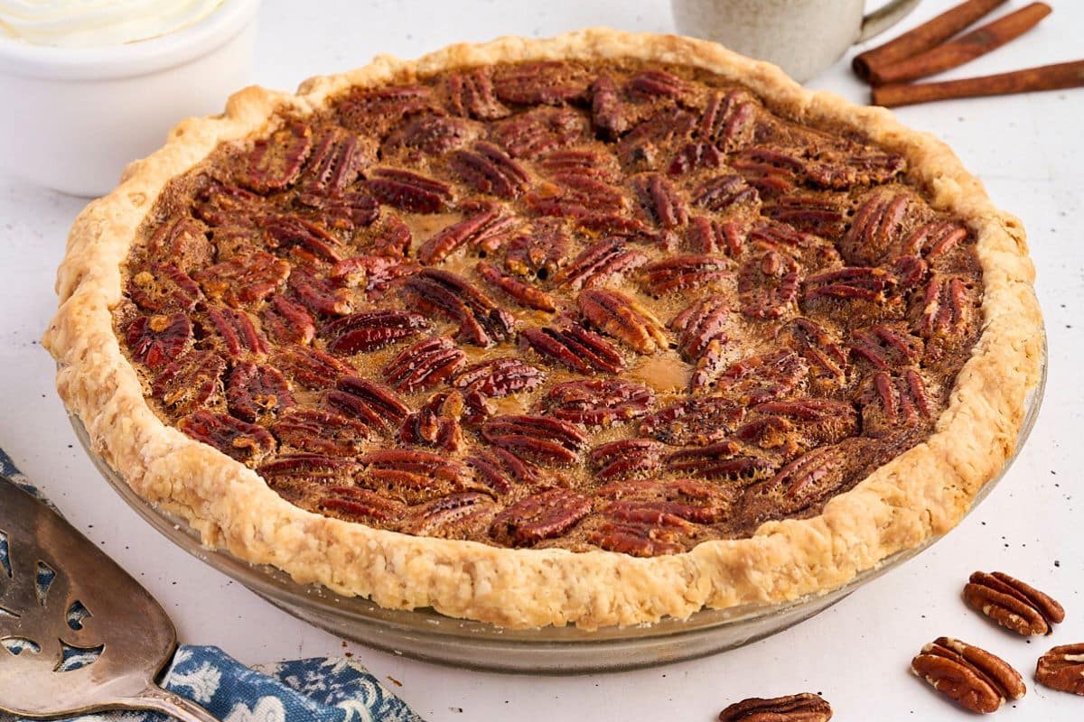 A pecan pie with a golden crust in a glass pie dish, surrounded by pecans and cinnamon sticks.