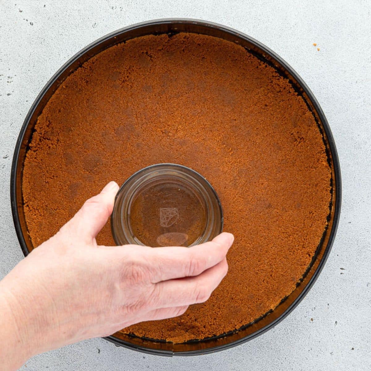 A hand presses a glass into a springform pan with a cheesecake crust.