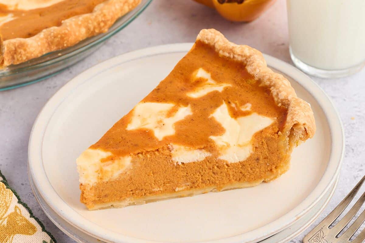 A slice of pumpkin pie with cream swirls on a white plate, next to a glass of milk.