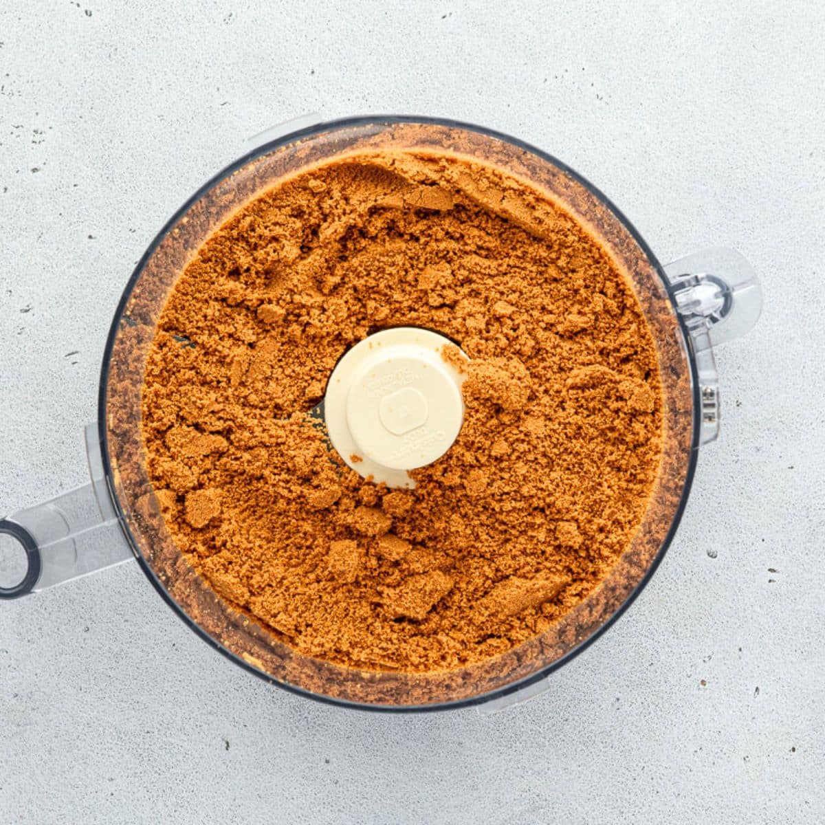 Ground cookie crumbs in a food processor bowl, viewed from above on a light gray surface.