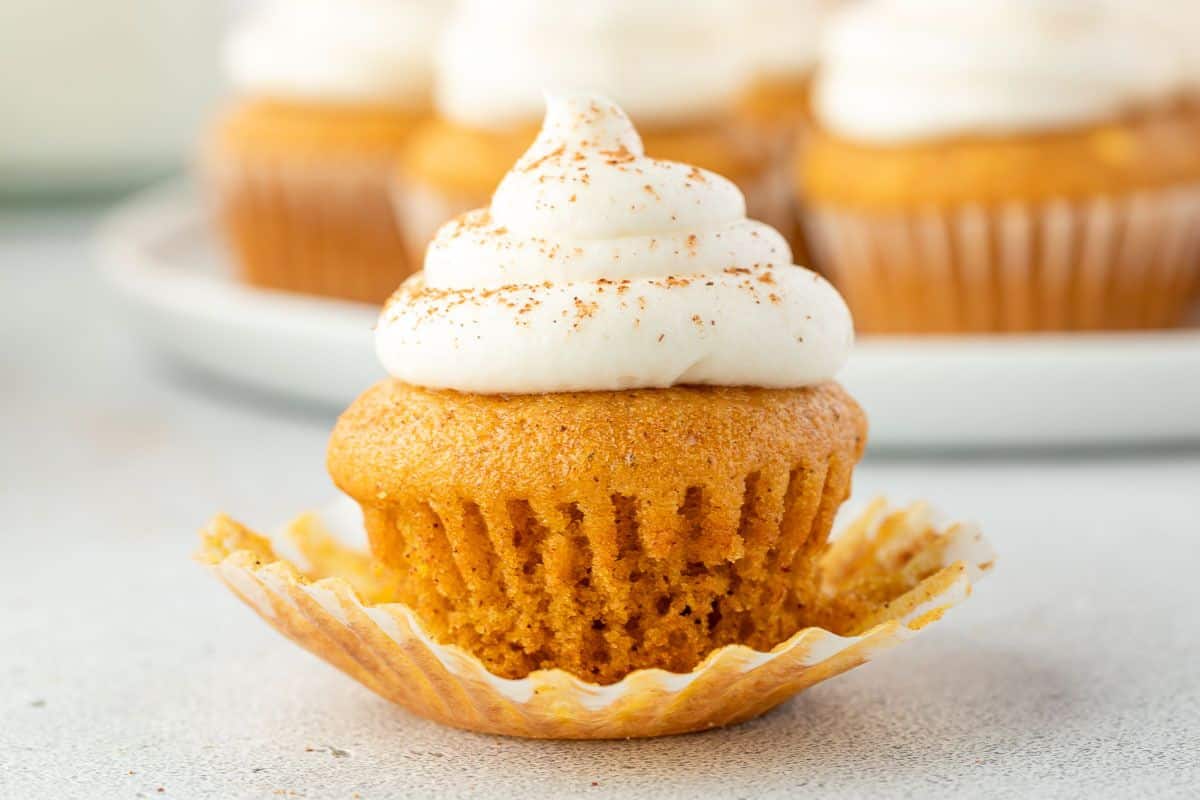 A pumpkin cupcake with white frosting and a sprinkle of cinnamon, with its wrapper partially peeled off.