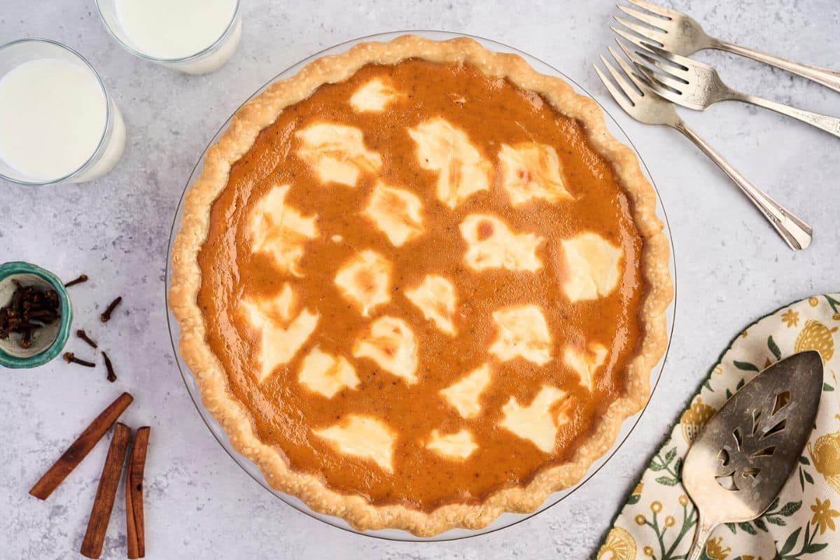 Pumpkin pie with cream cheese swirls, surrounded by forks, pie server, milk, spices, and a napkin.