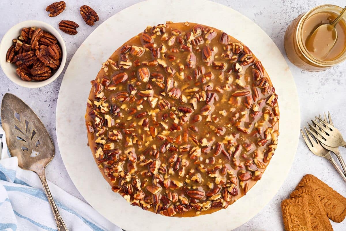 Round pecan caramel cake on a white platter, with utensils, pecans, and caramel sauce beside it.