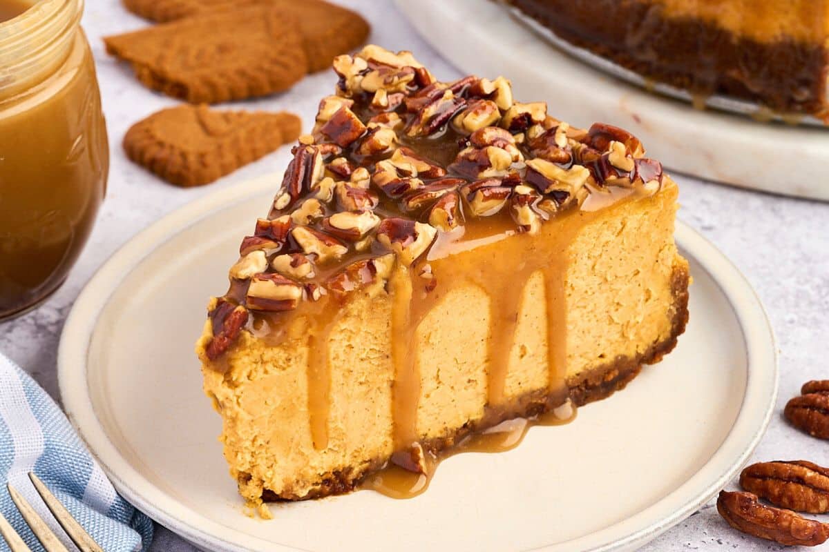 A slice of cheesecake topped with pecans and caramel sauce on a plate, with cookies in the background.