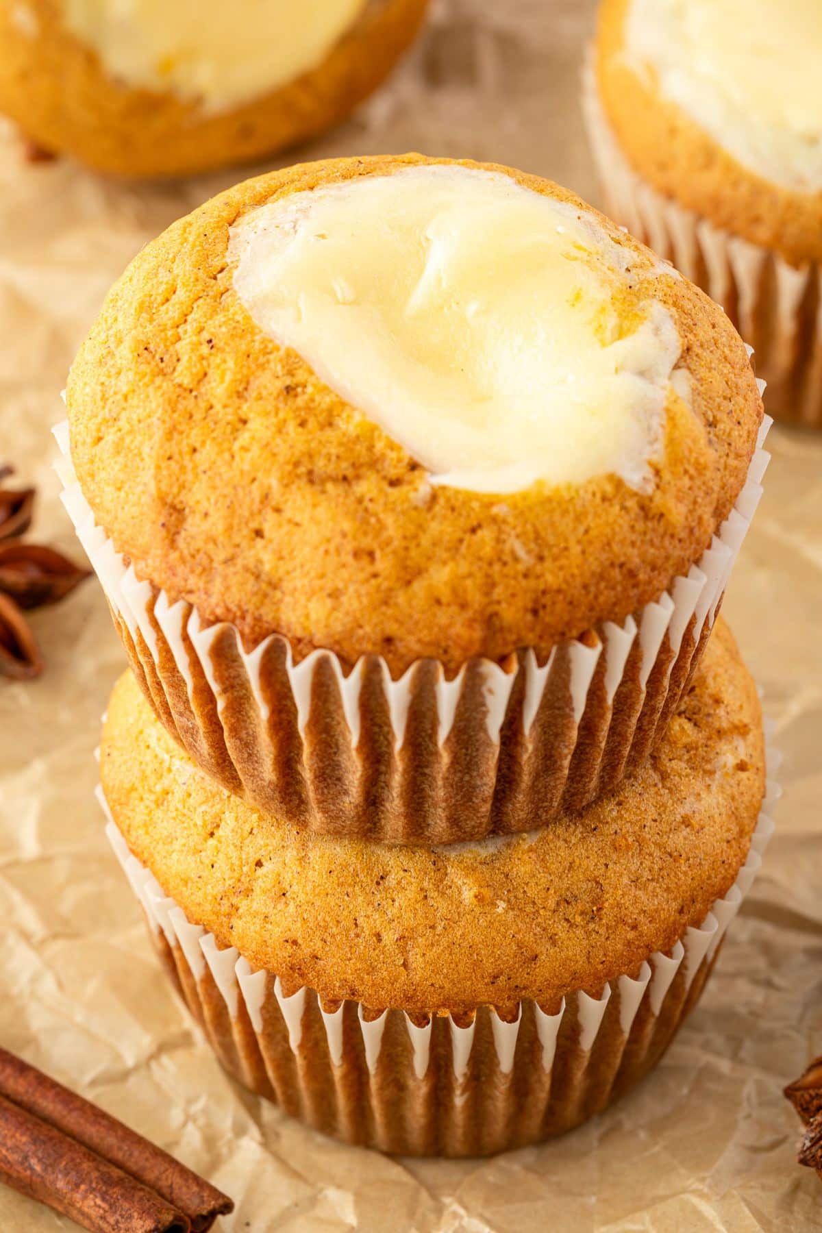 Two pumpkin muffins with creamy centers stacked on parchment paper, with cinnamon sticks in the background.