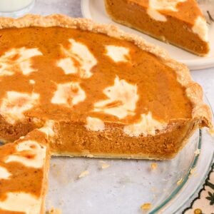 Pumpkin pie with a swirl of cream cheese, missing two slices, on a glass pie dish with one slice on a plate.