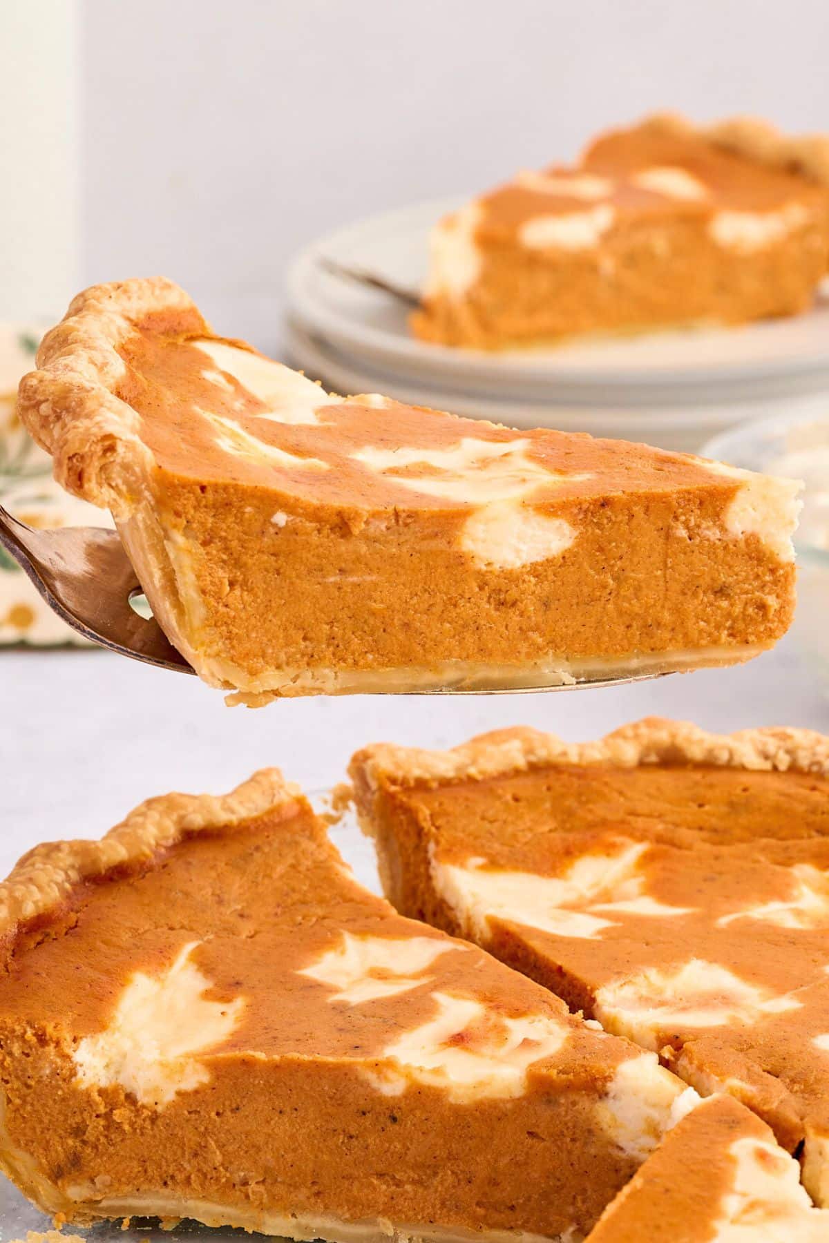 A slice of pumpkin pie with a creamy swirl pattern is being lifted from a whole pie.