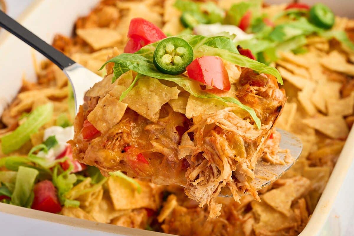 A spatula lifts a cheesy chicken casserole topped with chips, lettuce, tomato, and sliced jalapeño.