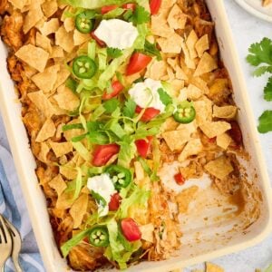 A baked casserole topped with crushed chips, lettuce, tomato, jalapeño slices, cilantro, and dollops of sour cream.