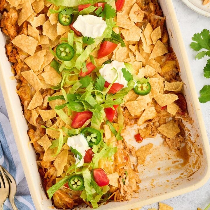 A baked casserole topped with crushed chips, lettuce, tomato, jalapeño slices, cilantro, and dollops of sour cream.
