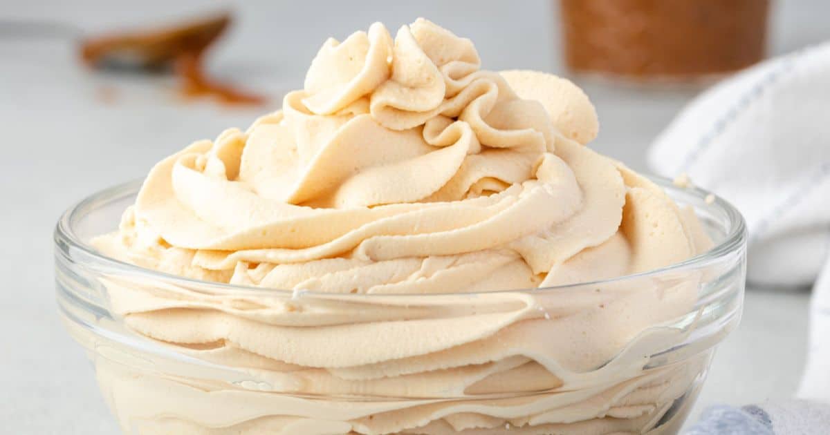 A glass bowl filled with swirled, creamy light brown whipped frosting on a light surface.
