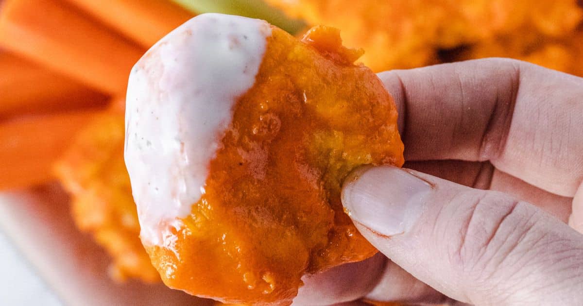 A hand holding a buffalo cauliflower wing dipped in ranch sauce, with carrot sticks in the background.