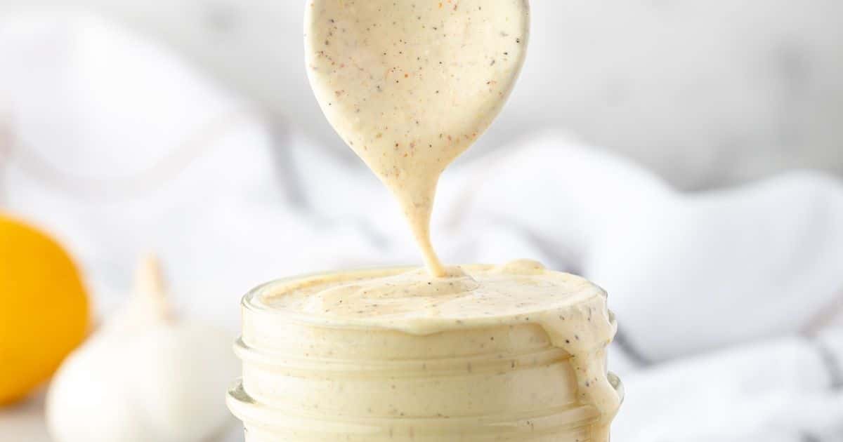 Spoon drizzling creamy, seasoned dressing into a glass jar with a blurred lemon and garlic in the background.