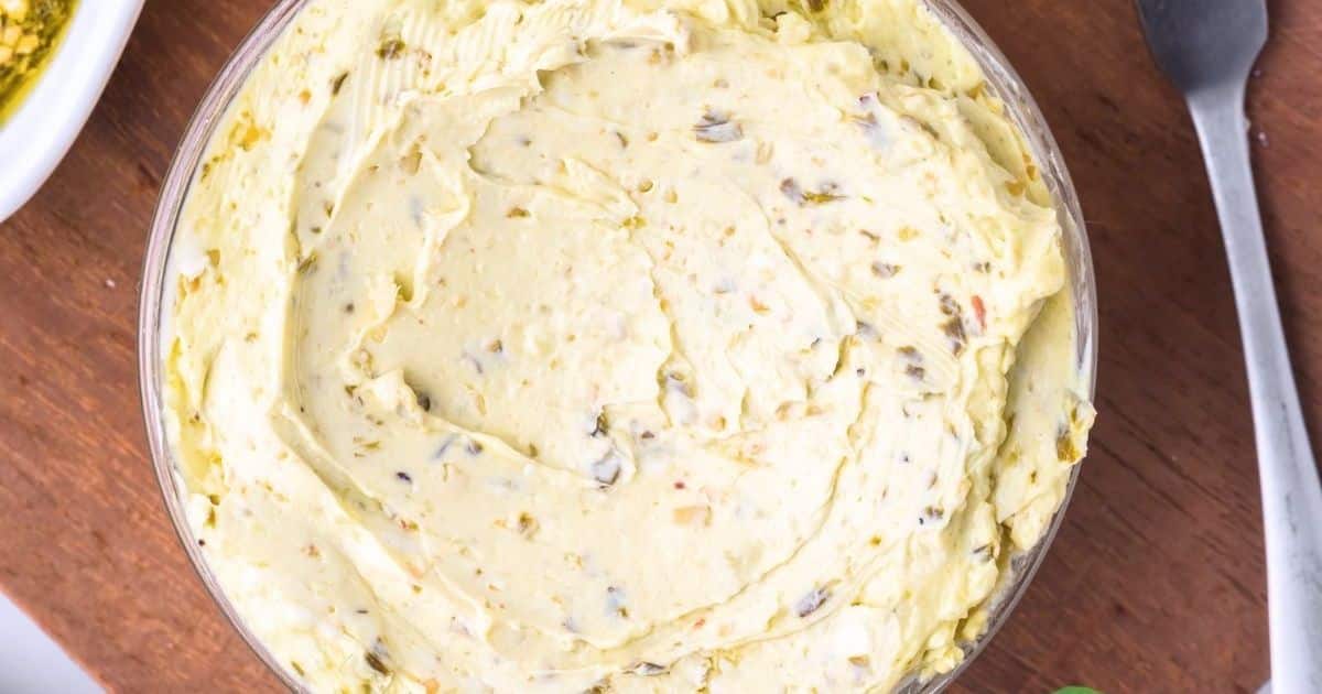 A bowl of creamy, herb-speckled butter on a wooden surface with a spoon beside it.