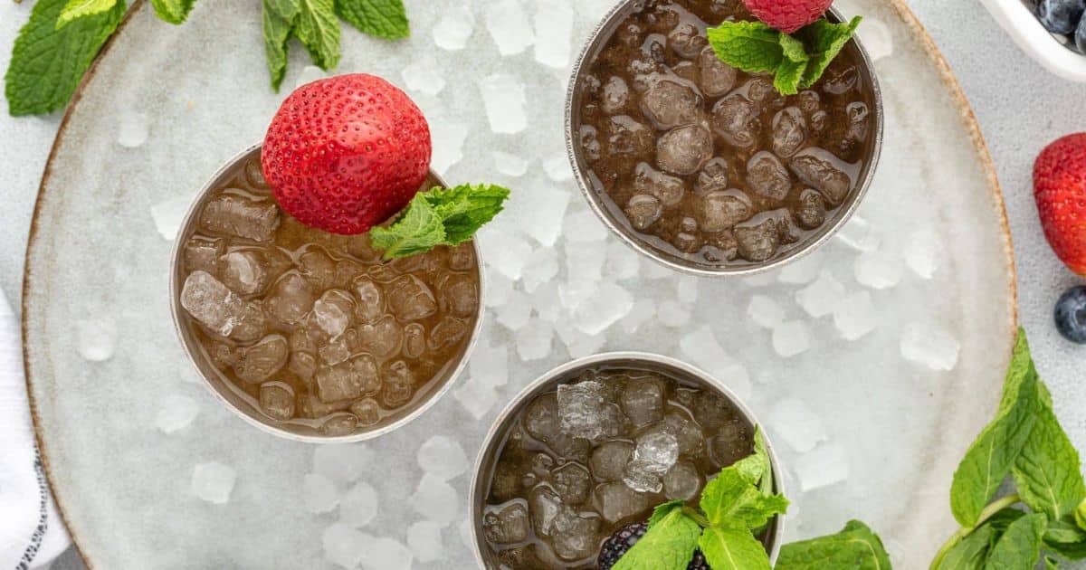 Three iced drinks garnished with mint and berries on a plate with crushed ice and scattered mint leaves.