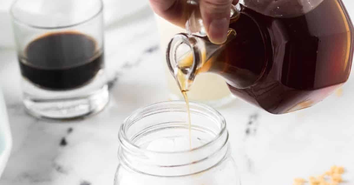 A hand pours syrup from a glass pitcher into a mason jar on a white surface, with drinks in the background.