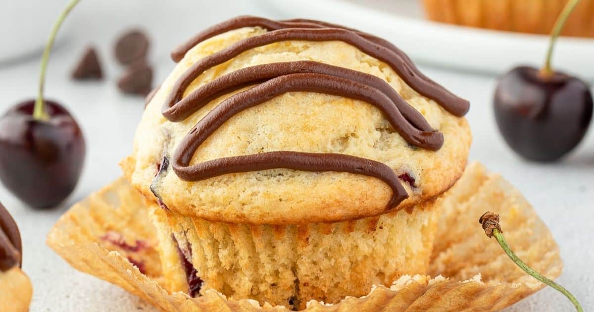 A cherry muffin with chocolate drizzle, unwrapped from its paper liner, sits near fresh cherries.