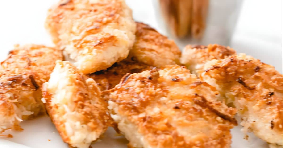 Golden brown coconut-crusted chicken tenders arranged on a white plate with dipping sauce in the background.