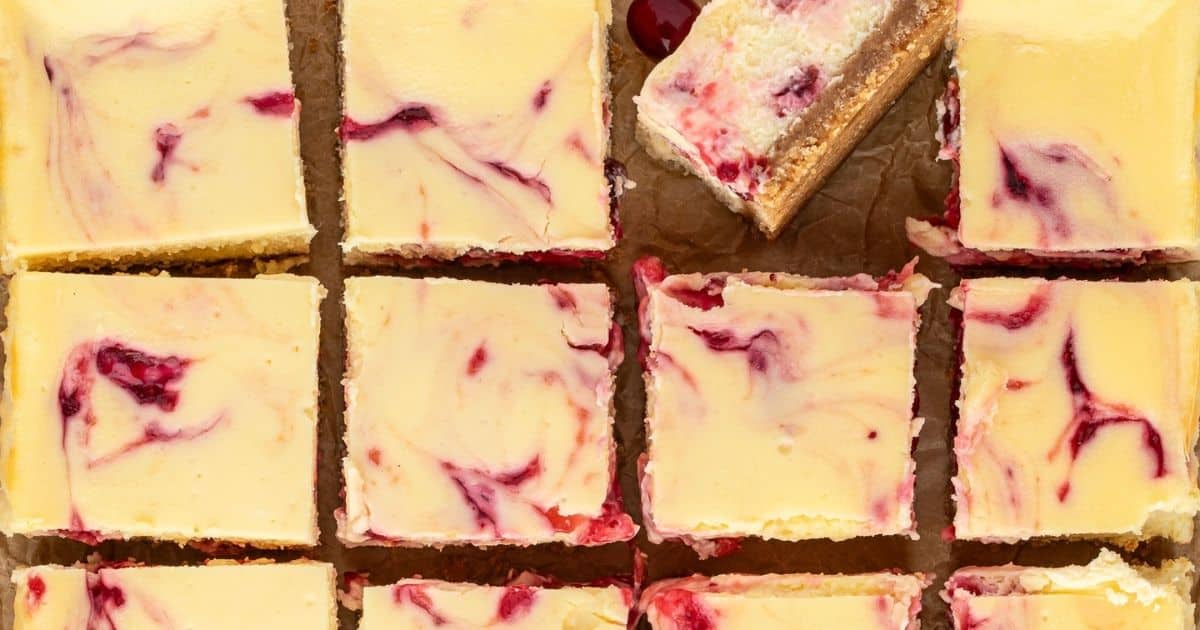 Squares of cheesecake with raspberry swirls arranged in a grid on parchment paper, one piece partially removed.