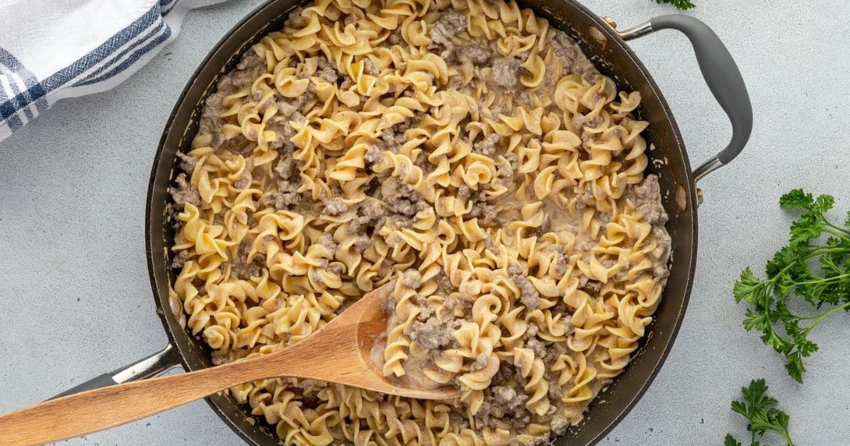 A skillet of creamy beef and noodle casserole with a wooden spoon, garnished with parsley on the side.