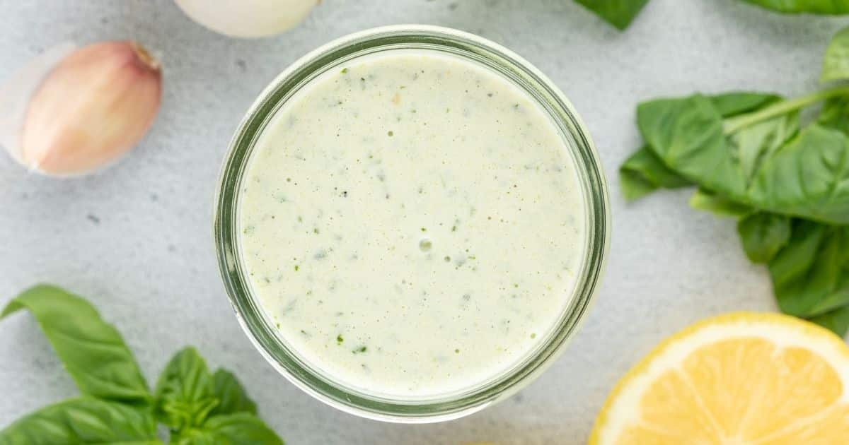 A jar of creamy green sauce surrounded by fresh basil, lemon, garlic, and onion on a light surface.