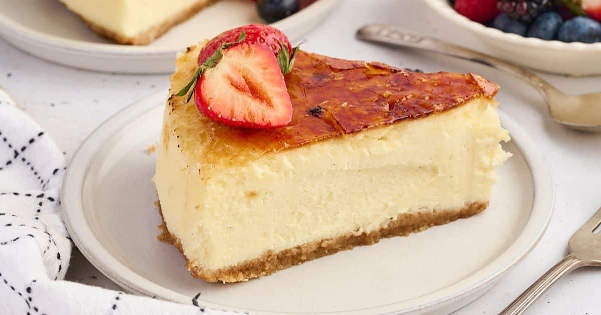 A slice of cheesecake with a caramelized top, garnished with a halved strawberry on a white plate.