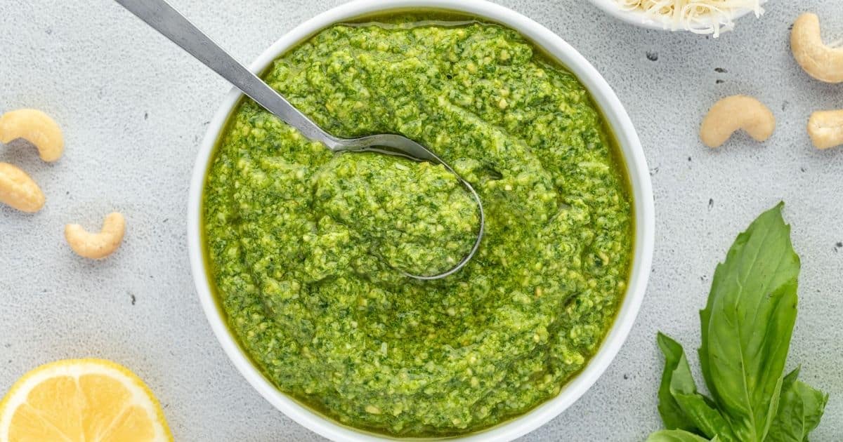 A bowl of green pesto sauce with a spoon, surrounded by cashews, basil leaves, and a lemon slice.