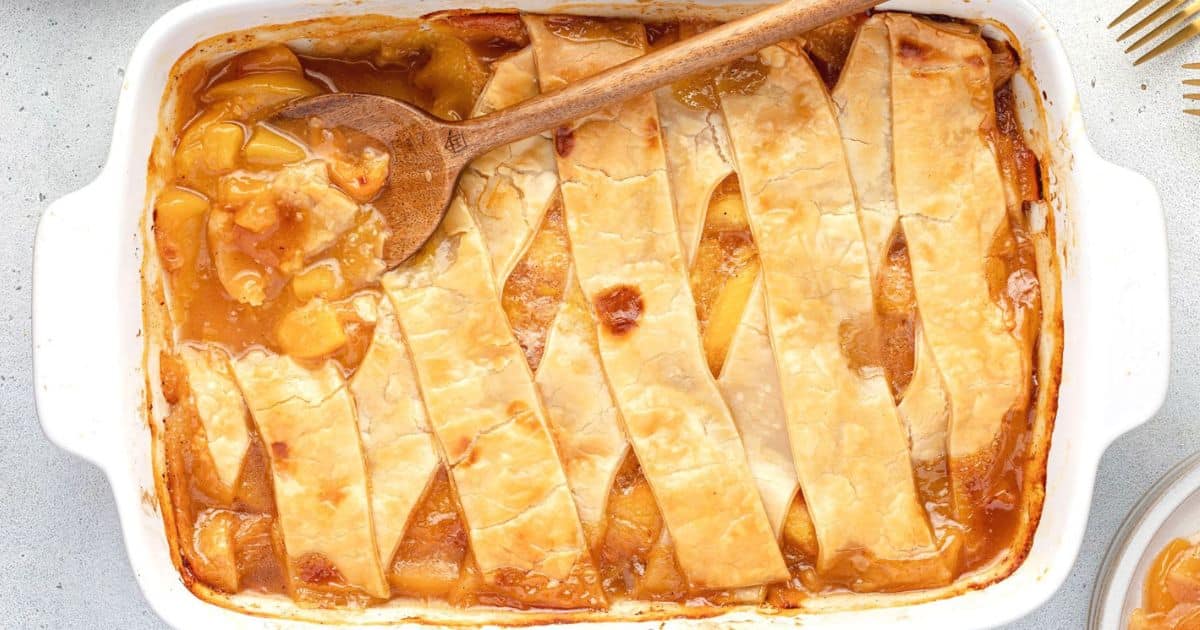 Rectangular dish of peach cobbler with a lattice crust, a wooden spoon scooping some from the corner.