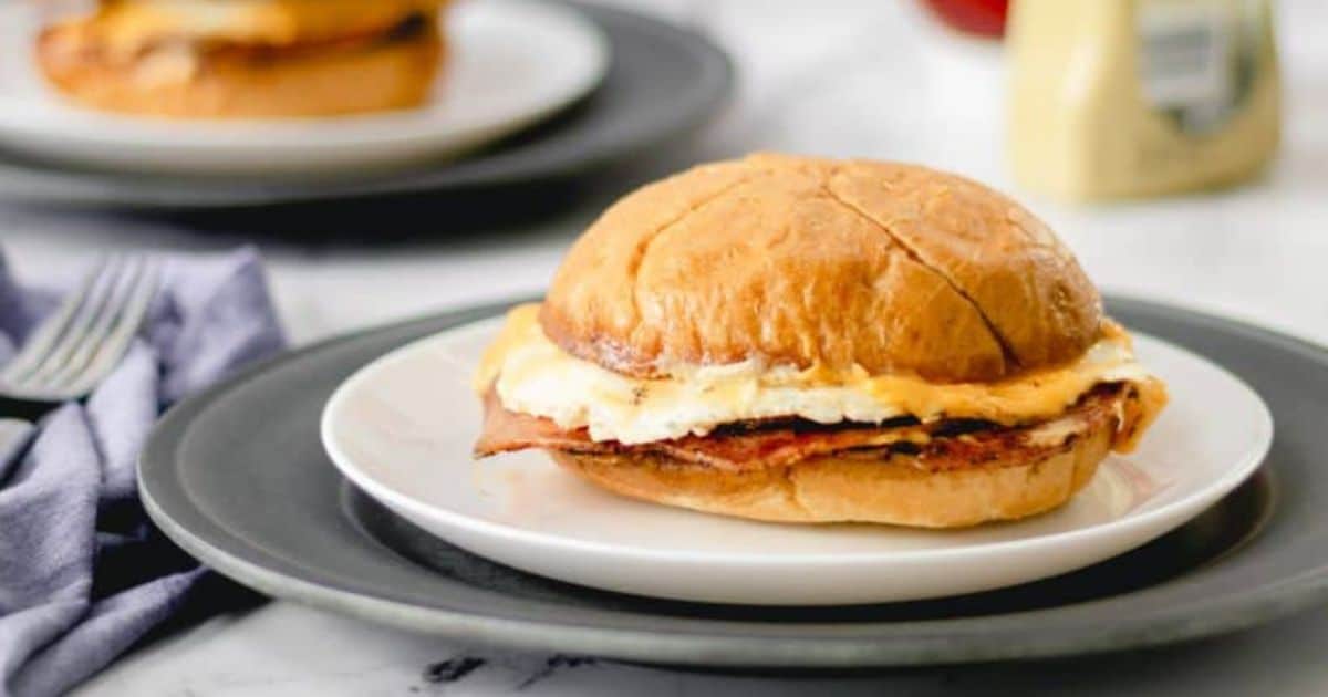 Breakfast sandwich with egg and bacon on a bun, served on a white plate with another plate in the background.